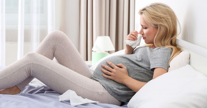 Pregnant woman with allergy sitting on bed at home; blog: ways to avoid getting sgick while pregnant