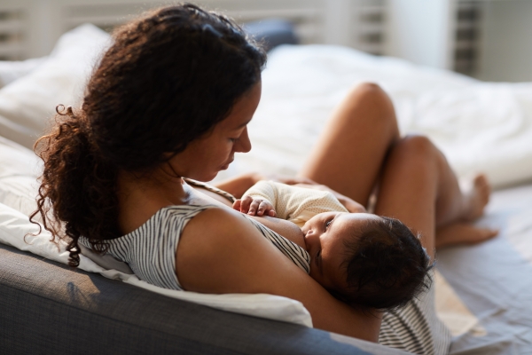 Peaceful loving young African mother sitting on bed and leaning on headboard while feeding baby with breast; blog: Breastfeeding Tips For New Moms