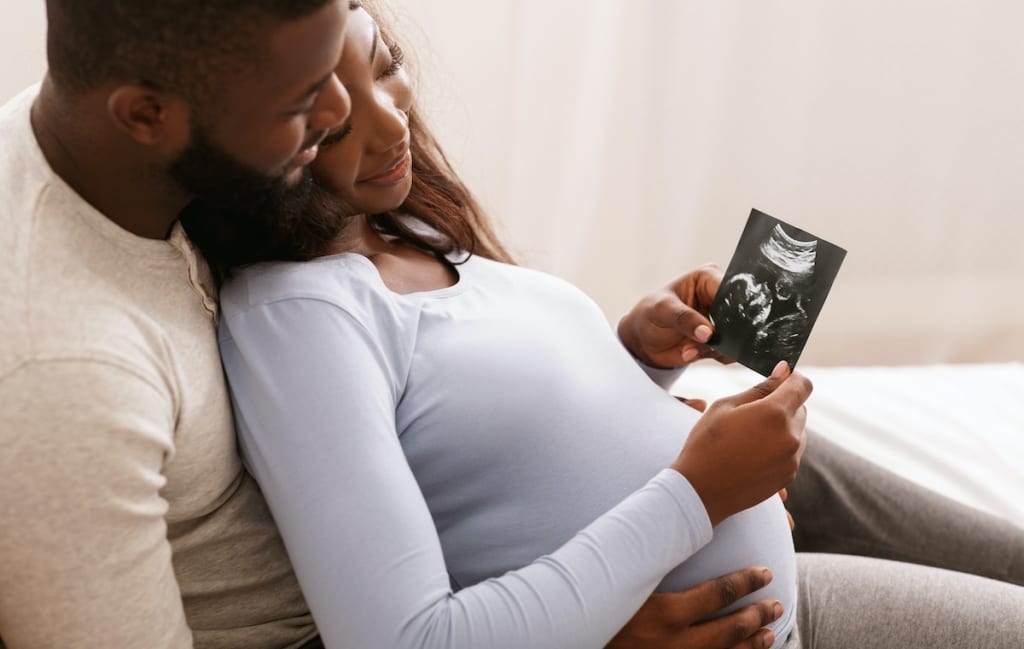 Relaxed Black couple watching baby ultrasound photo, lying in bed at home; Pregnancy: Fetal Ultrasound